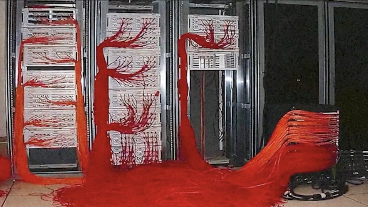 A mass of red wires on the floor and wired up to servers in a server room, low quality image.
