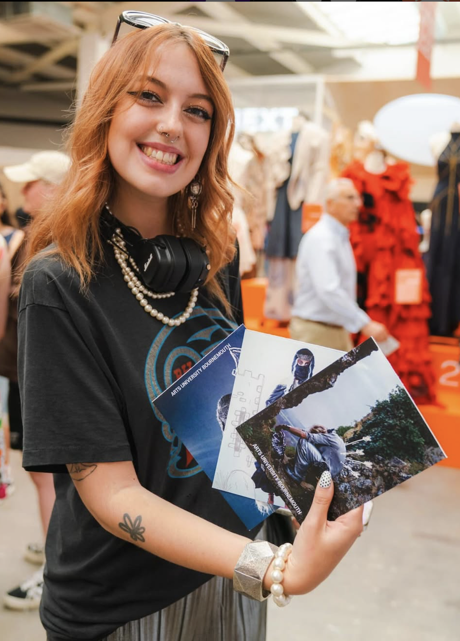 BA (Hons) Fashion Communication student L B Wyatt poses for a photo while holding a vinyl record in a busy shop, wearing dark T-shirt and pearl necklace.