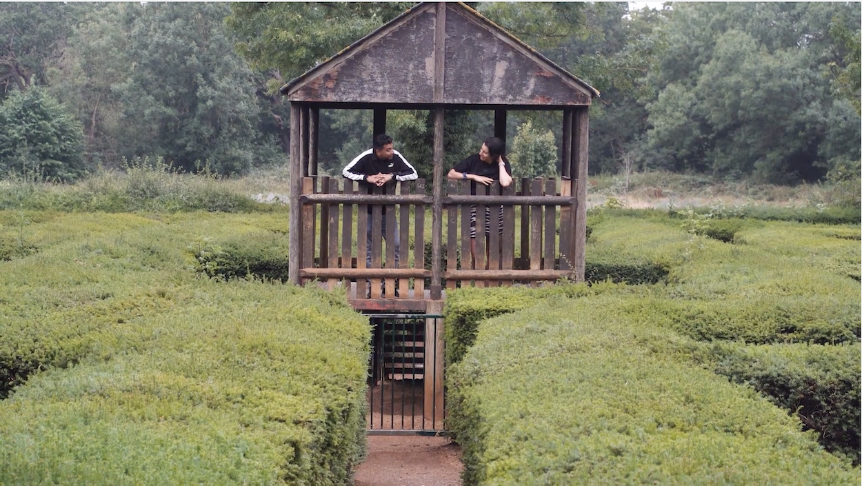 Wooden gazebo on stilts in centre of hedge maze, with two people visible inside the raised structure surrounded by green hedgerows.