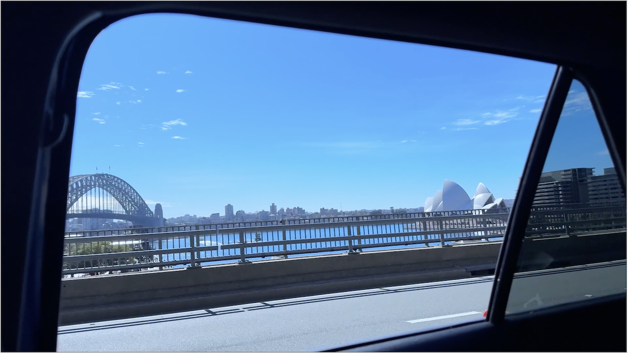 View through car window showing Sydney Harbour Bridge and Opera House across blue water under clear sky with white clouds.