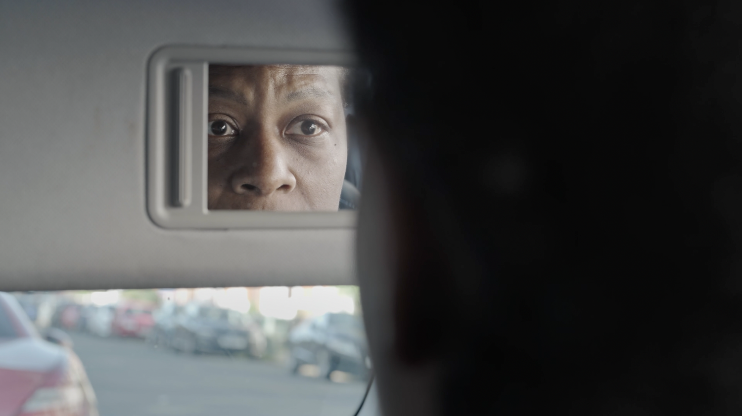 Woman's face reflected in rectangular car sun visor mirror, blurred road visible through window below.