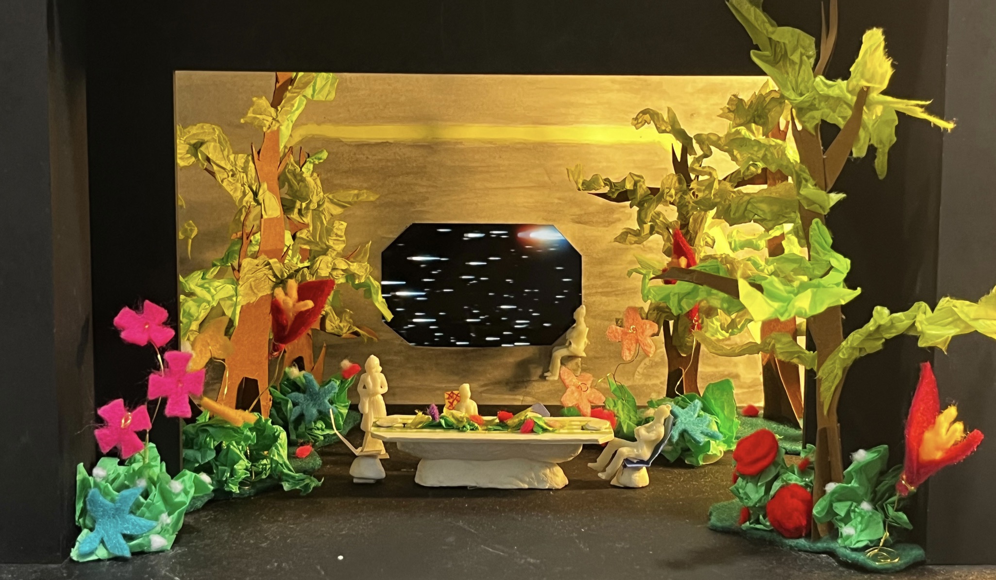 A miniature diorama shows two figures seated at a small table between painted trees and colourful flowers, with a dark octagonal window-like opening in the background.