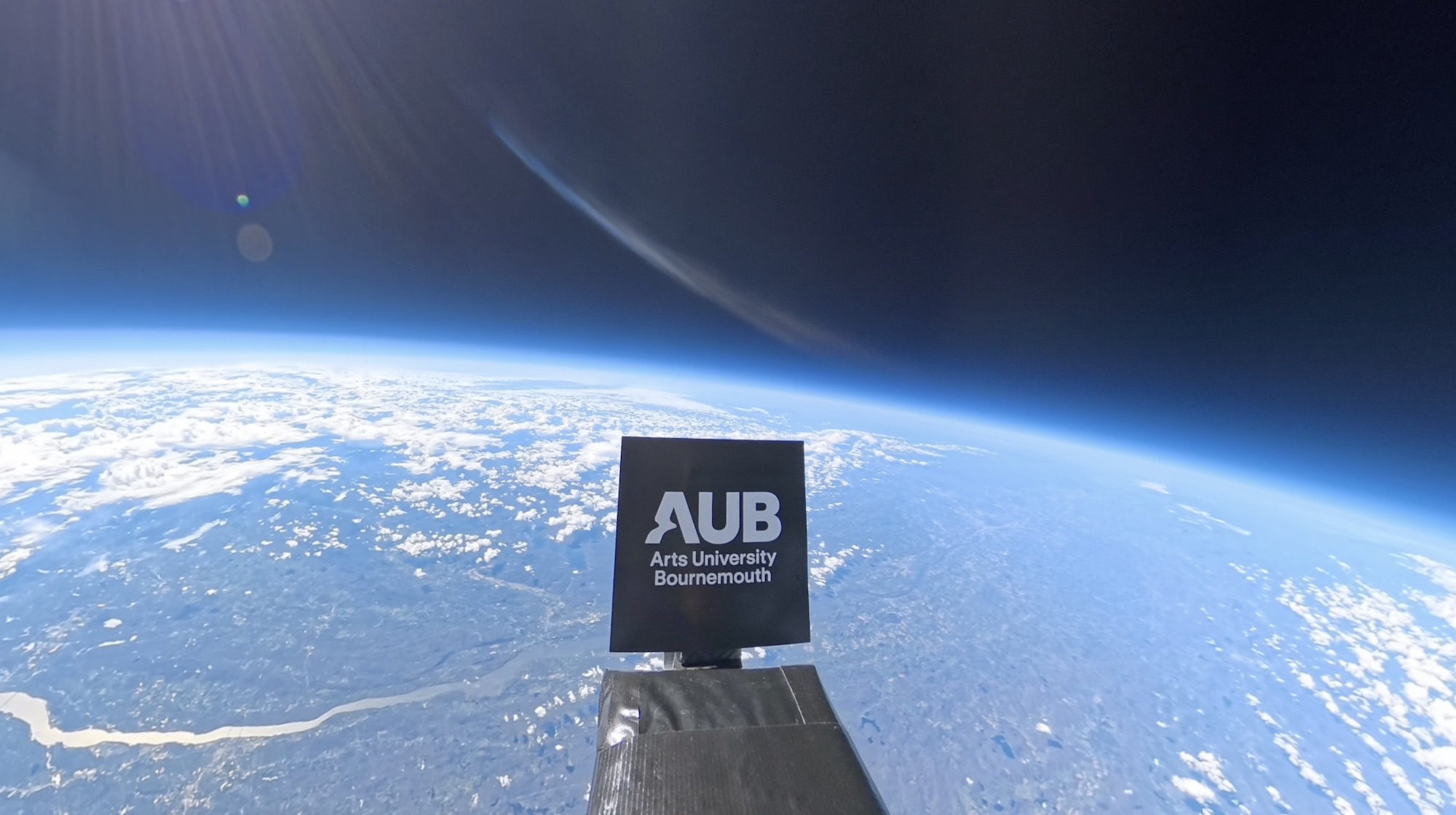 A black AUB Arts University Bournemouth sign mounted on a spacecraft, with Earth’s curved horizon and cloud cover in the background.