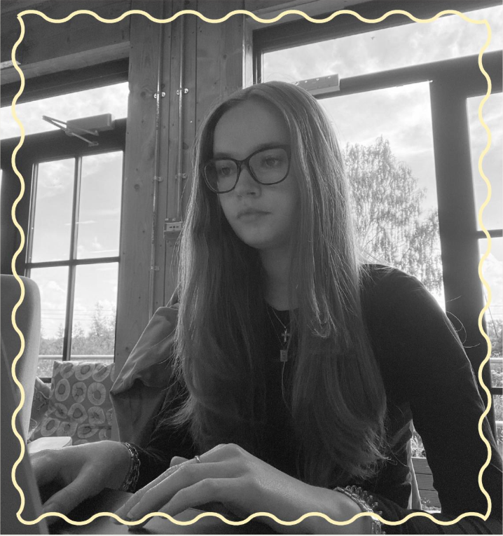A girl with long hair and glasses is sitting at a table, focusing on her laptop while typing. The background features large windows with natural light and trees visible outside, with a rustic wooden interior and patterned seating.