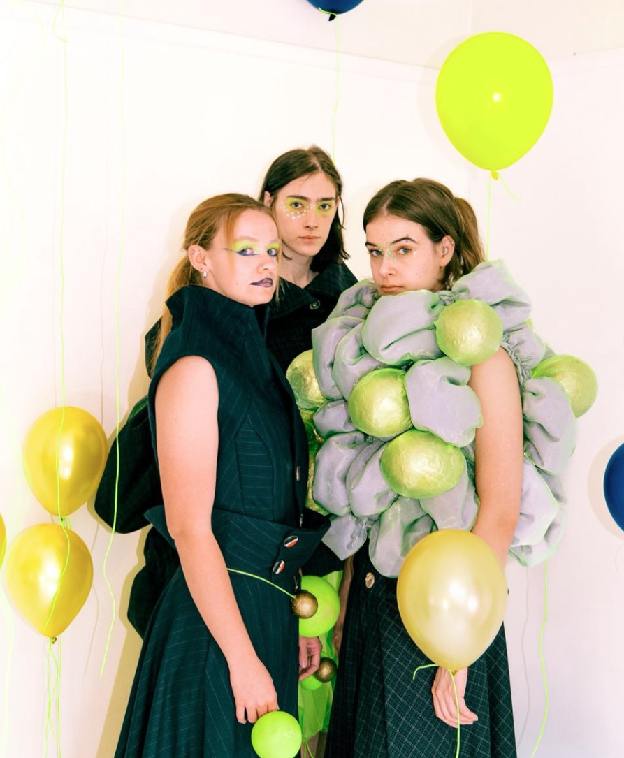 Three women stand together in a brightly lit room decorated with yellow balloons. The first woman, on the left, wears a navy dress and has a bold makeup look. The second, in the middle, wears a green and grey sculptural outfit, while the third, on the right, wears a white dress and also holds a balloon.