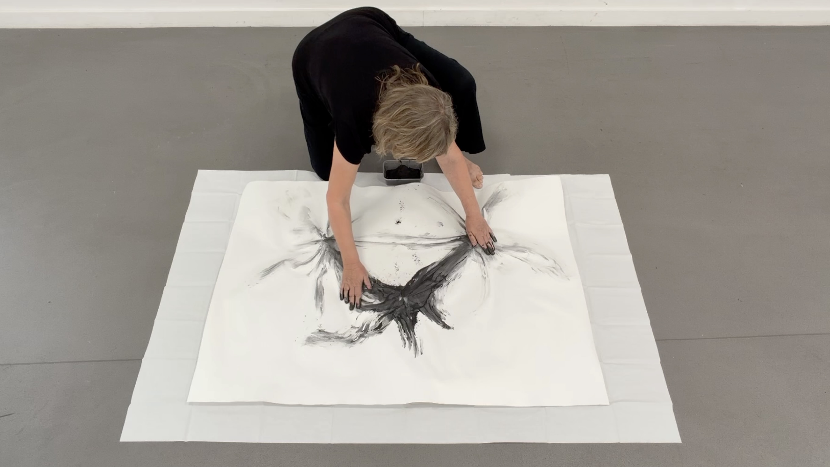 A woman bends over a large white sheet on the floor, creating a charcoal drawing with sweeping black marks in a plain room.