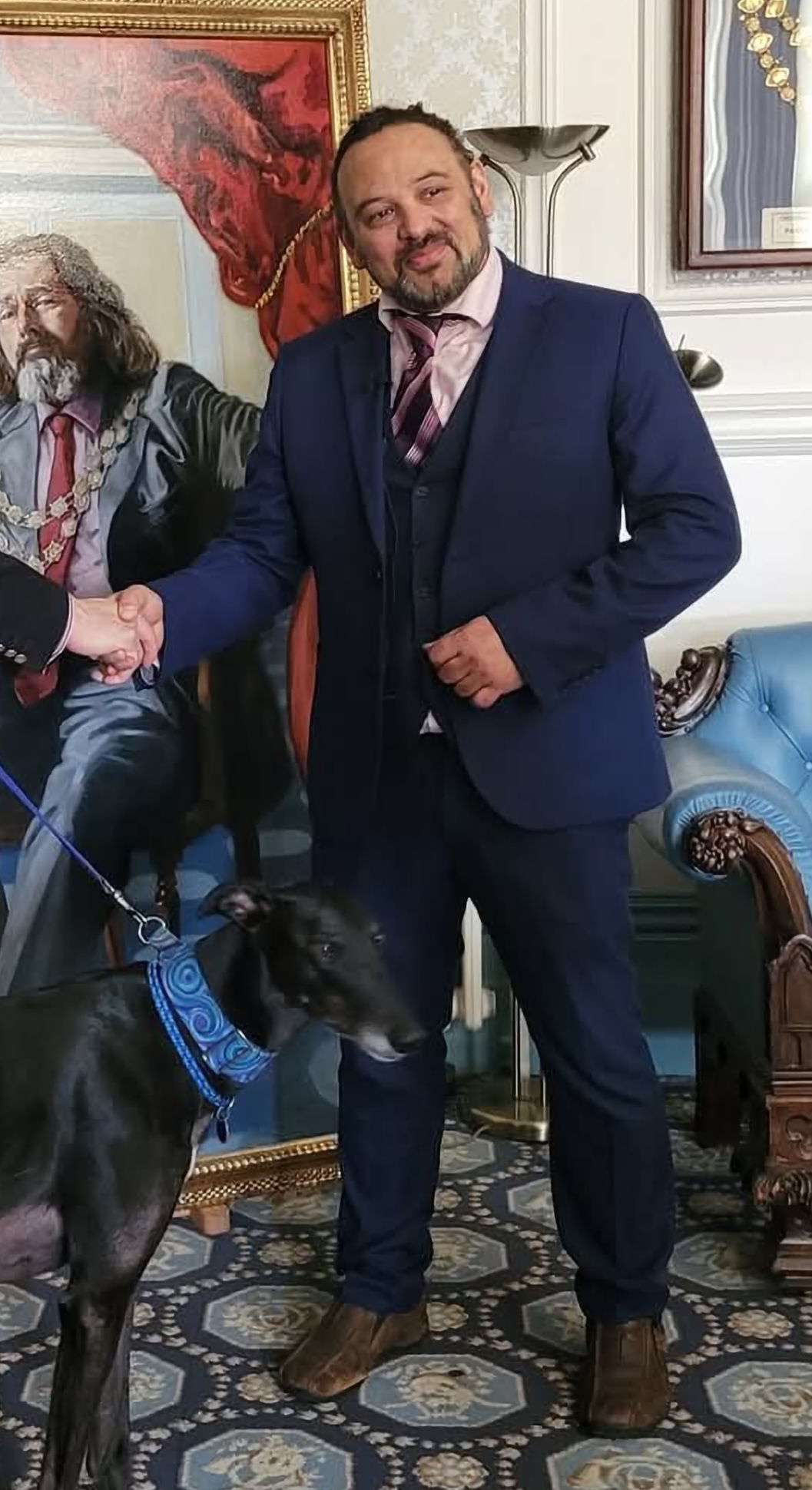 Man in navy suit with purple tie standing in ornate room with dark dog, framed paintings on white walls, decorative furniture.