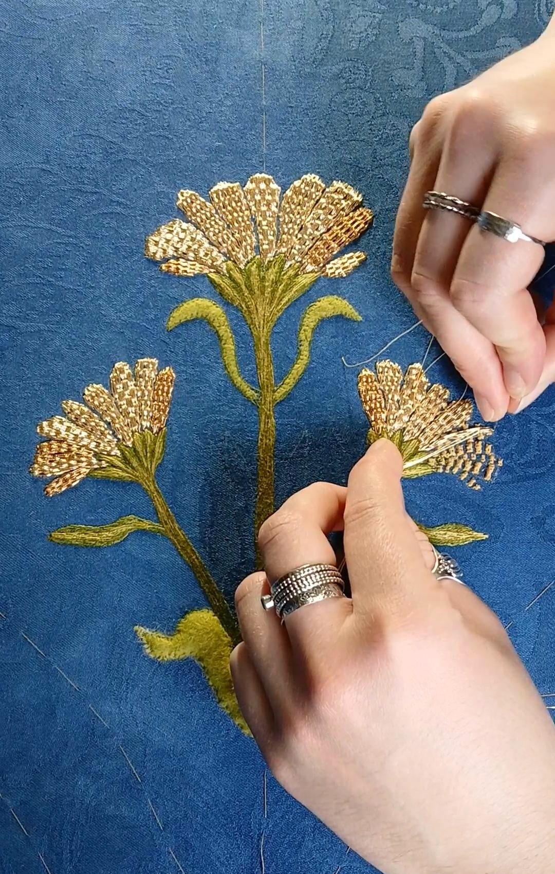 Hands embroidering gold thread flowers on blue fabric, wearing silver rings, detailed metallic floral pattern with green stems.