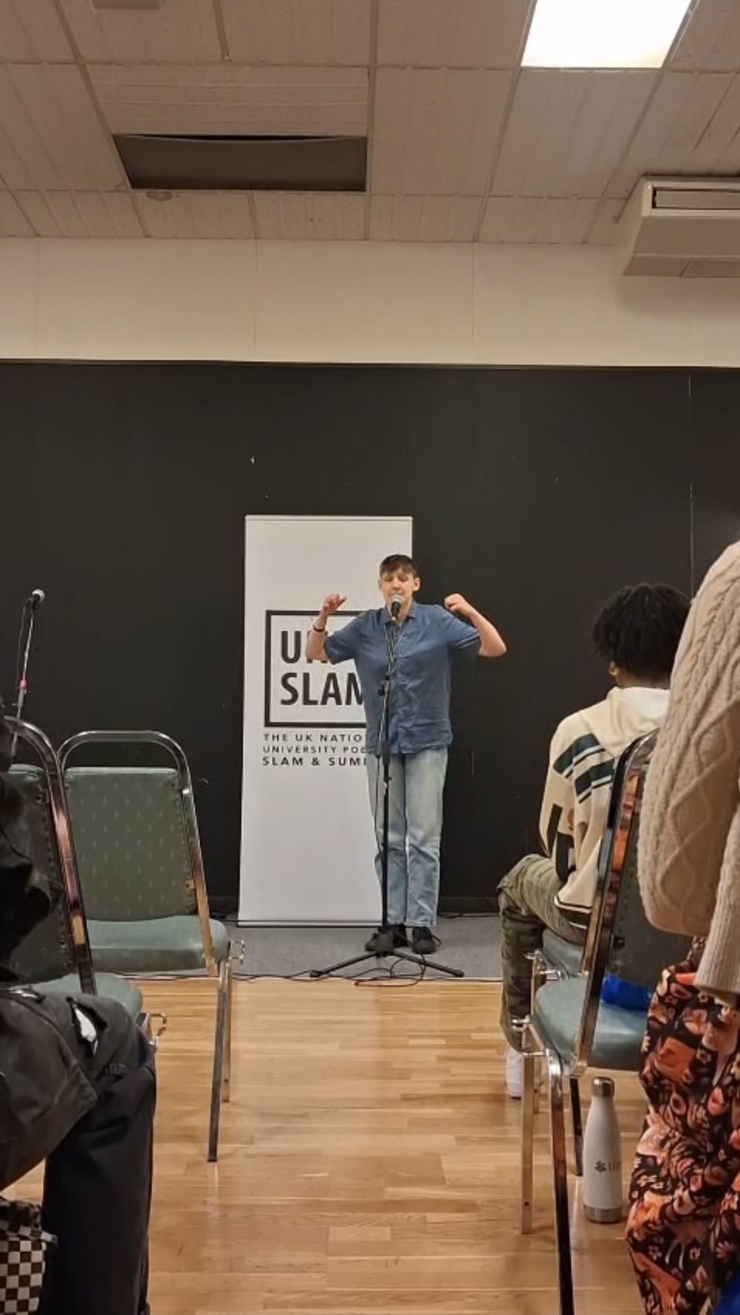Student performing to audience with mic, white UniSlam banner behind them.