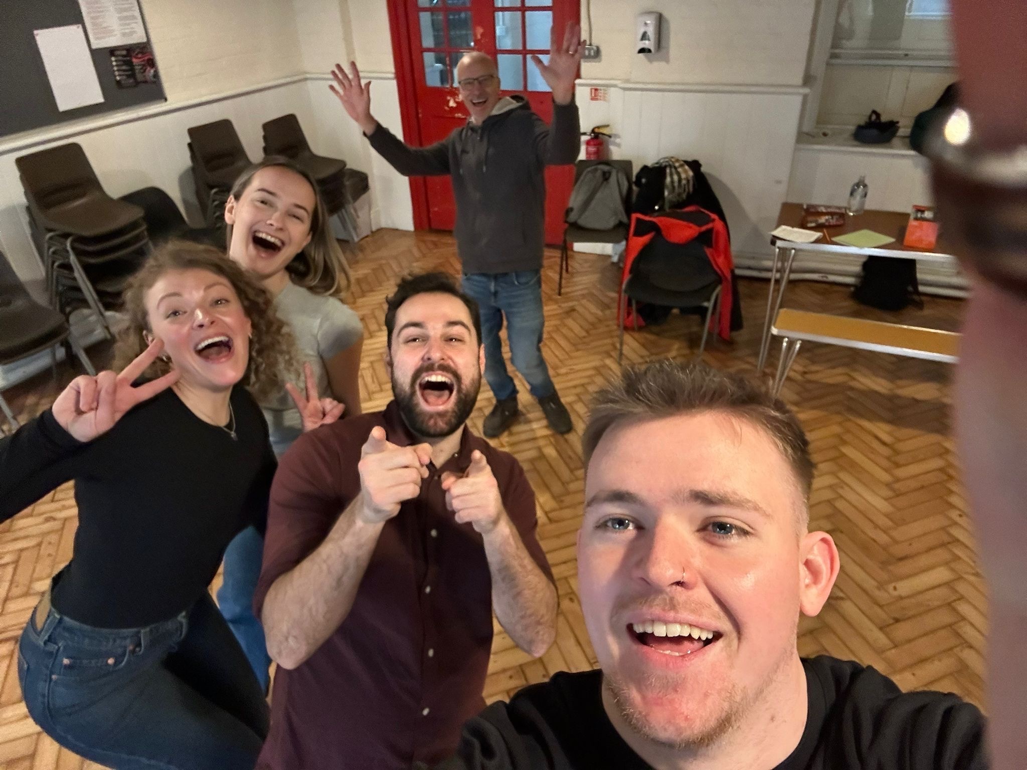 Five people taking selfie in rehearsal room, all smiling and gesturing enthusiastically. Man in foreground holds camera, others wave and make peace signs behind.