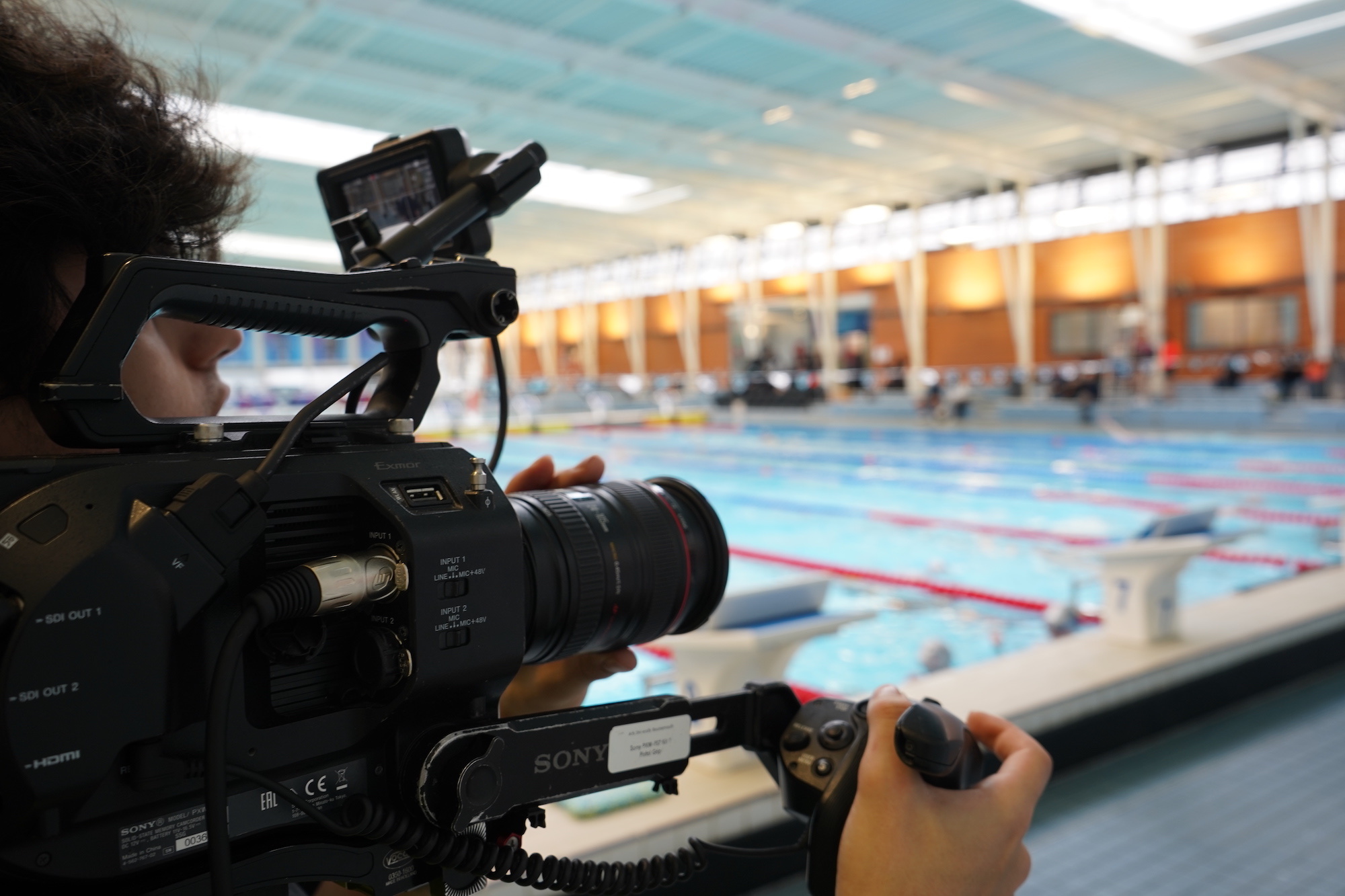 Film camera held by a crew member filming in an indoor swimming pool.