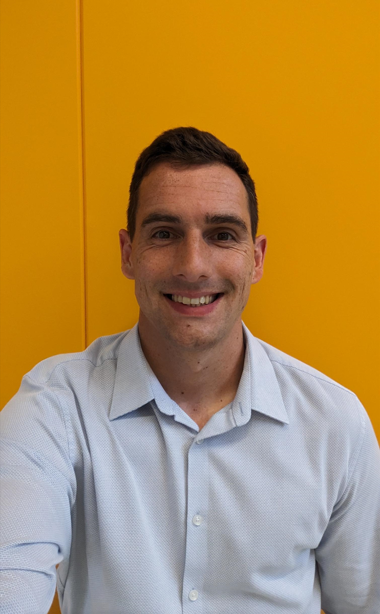 Man with short dark hair wearing light blue collared shirt, smiling at camera against bright yellow wall background.