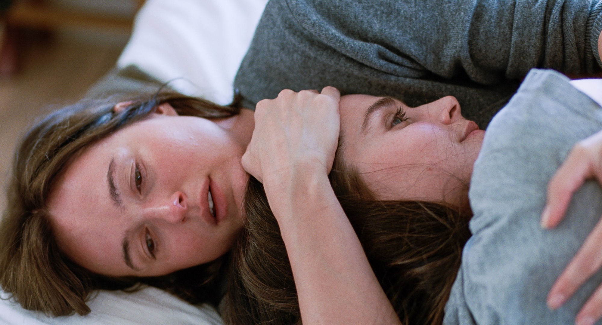 Two people lying close together on white bedding, woman with brown hair in foreground, man in grey top behind her.
