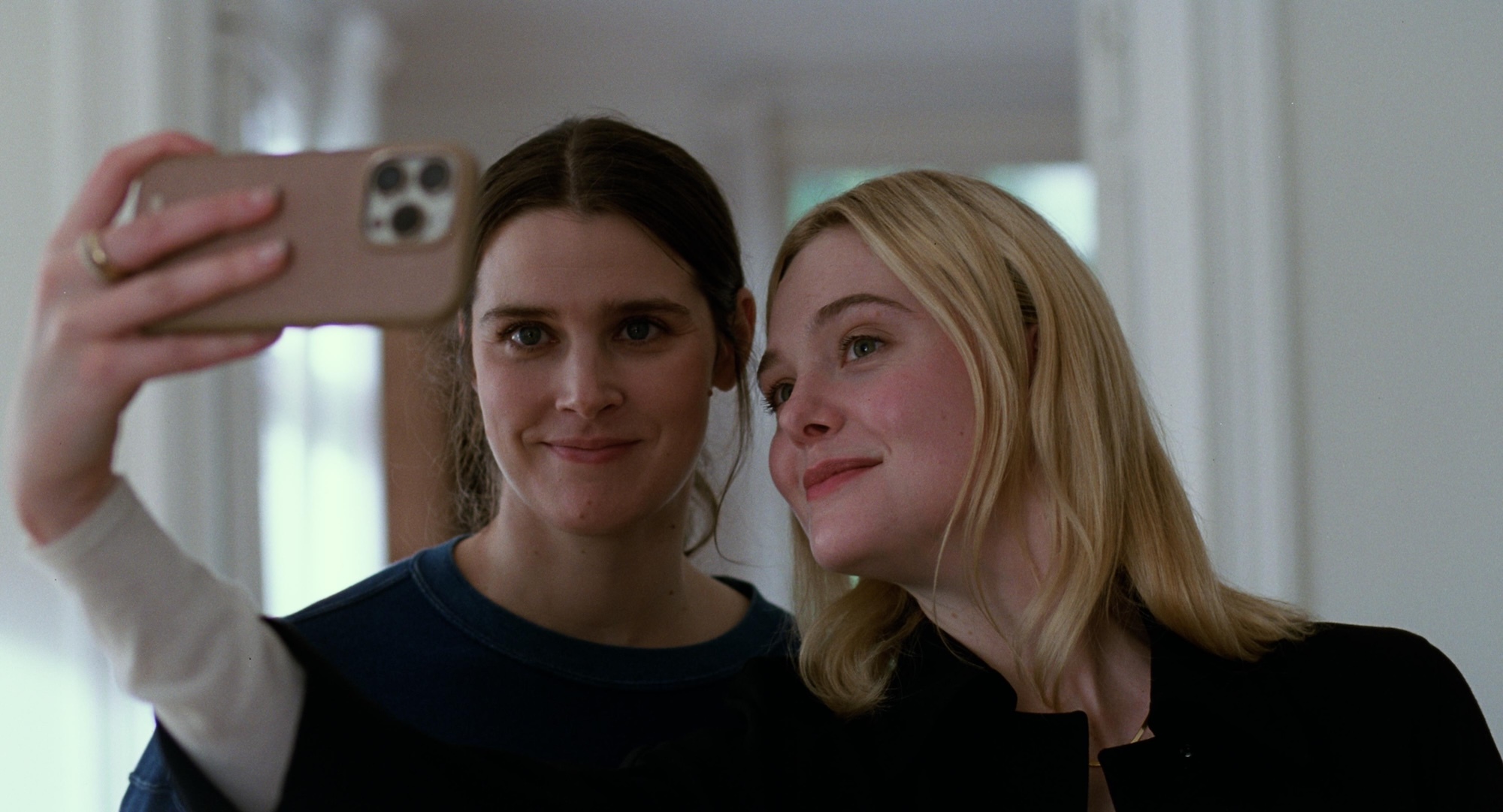 Two women taking a selfie with a mobile phone indoors, one with dark hair in black top, one blonde in black jumper.