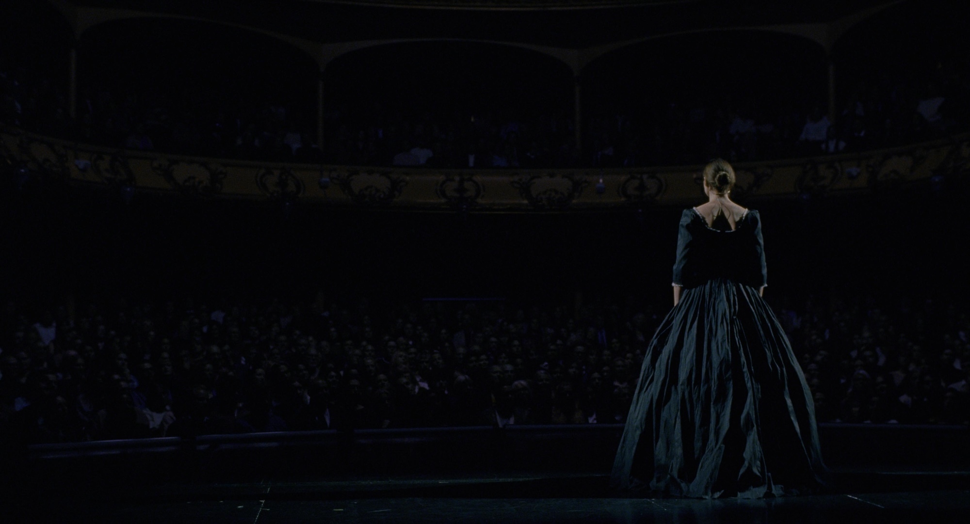 R Reinsve portraying character with back to camera, standing on stage in period costume, facing out to a dark auditorium. Still from "Sentimental Value" (2025)