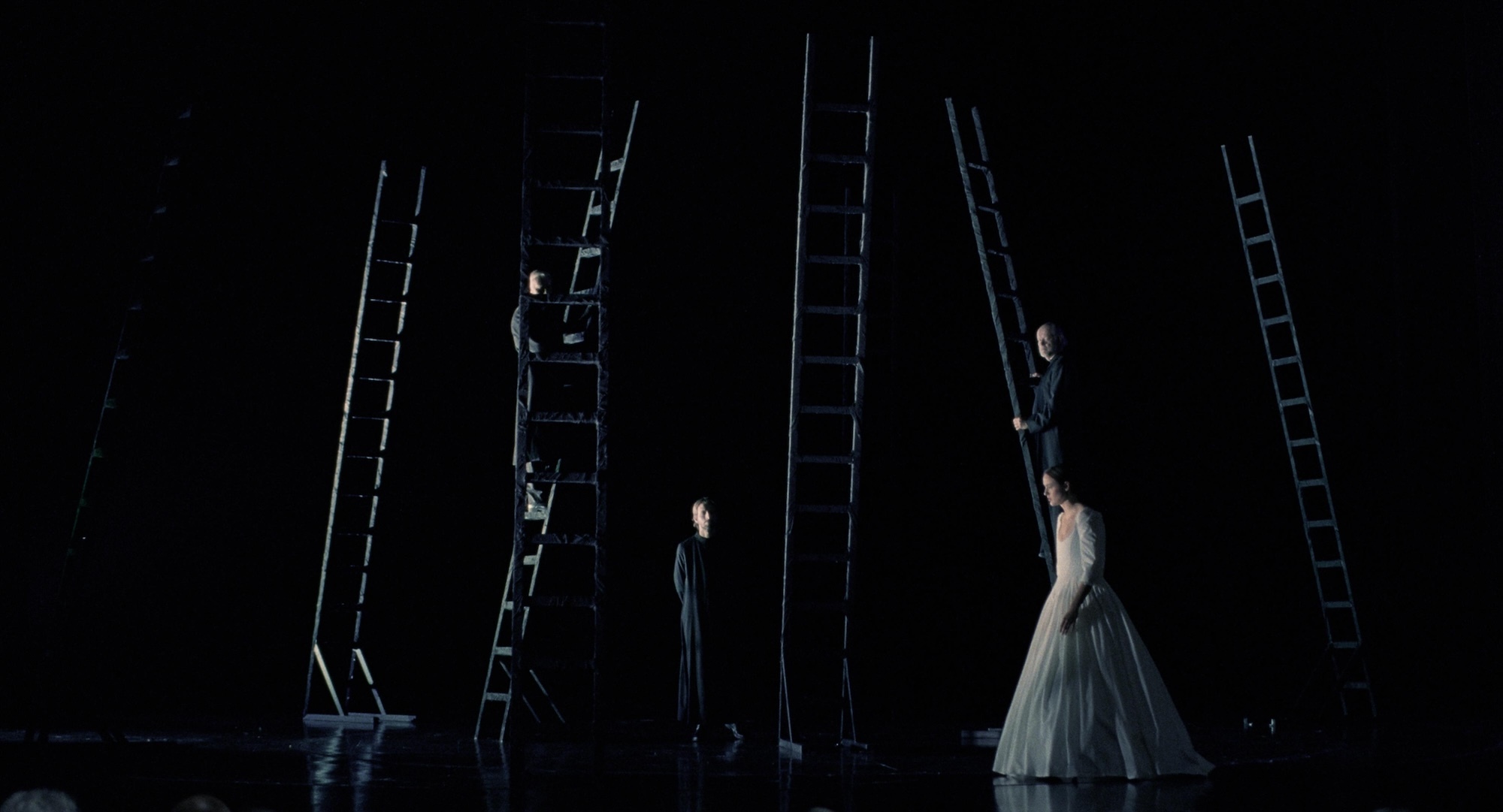 Dark stage with multiple tall ladders and performers in white and dark clothing positioned at various levels against black background.