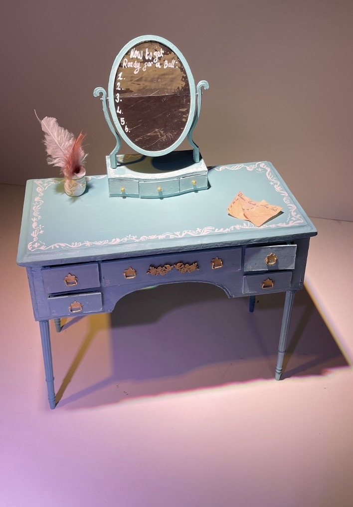Set design model of a light blue vanity table with a round mirror, pink feathered pens and old paper, stood in a studio with warm-pink lighting.