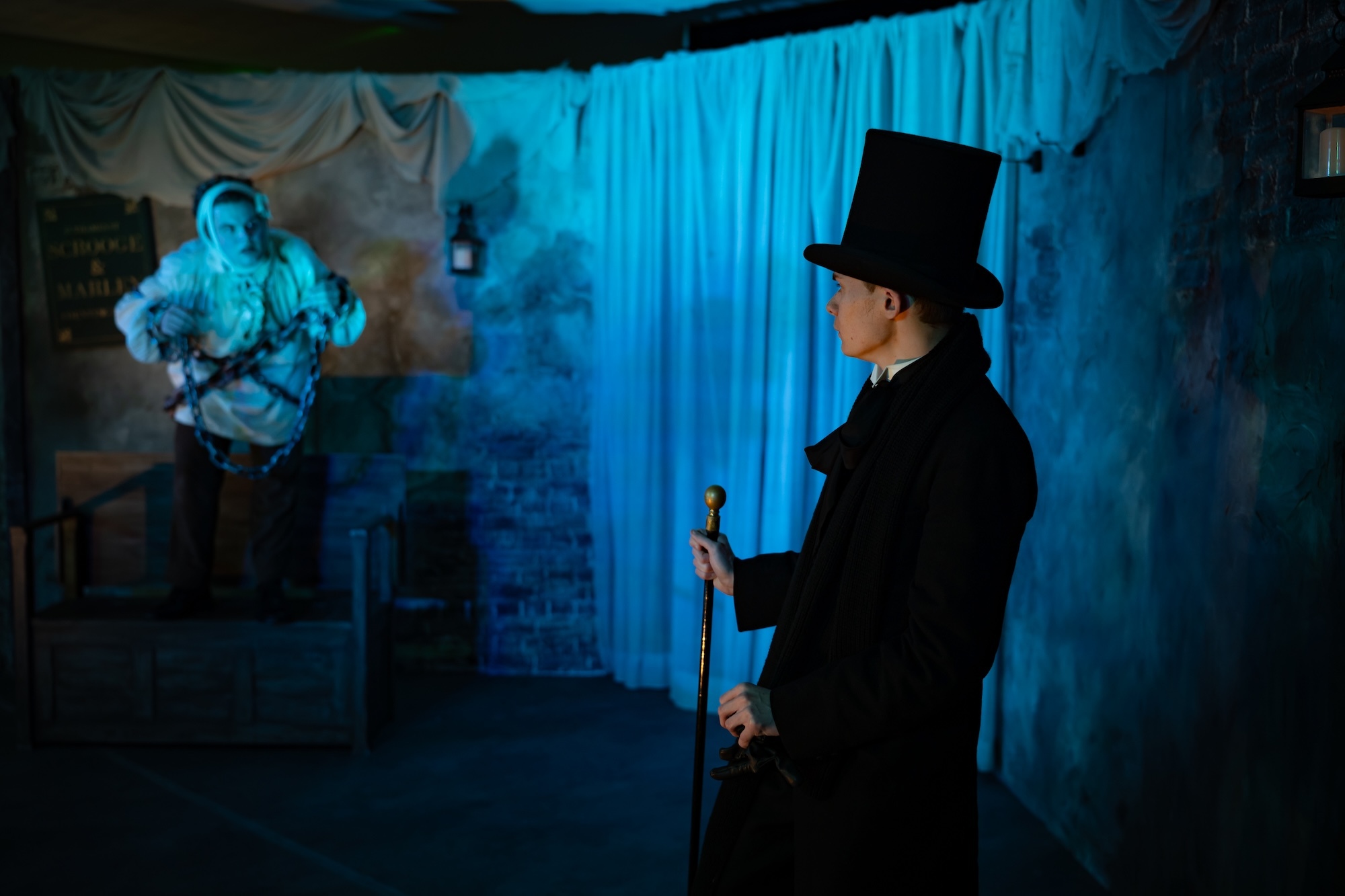 Man in top hat and dark coat holding cane faces ghostly blue-lit figure in smoky indoor setting with draped fabric backdrop.