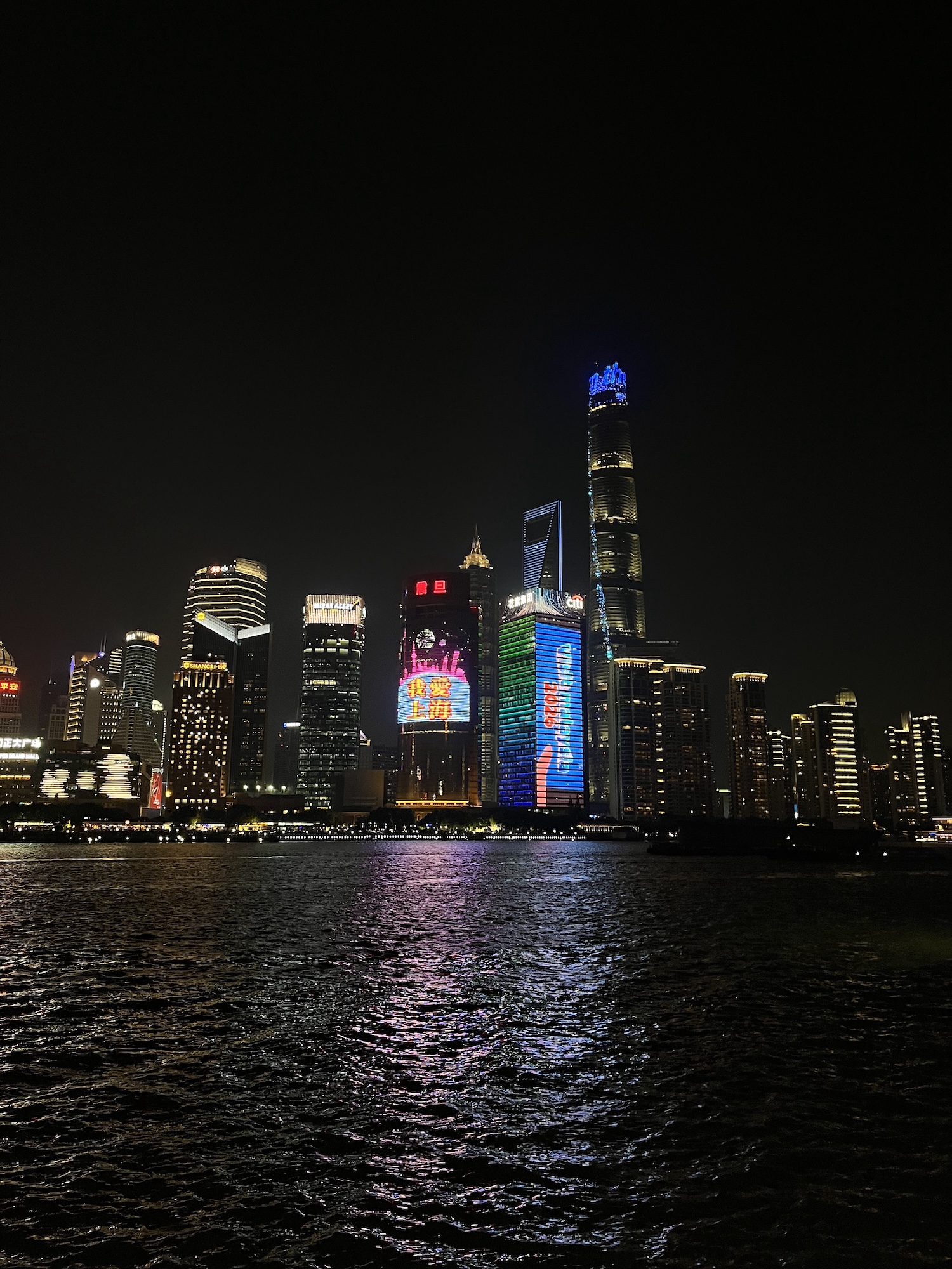 A night skyline of illuminated skyscrapers reflected on dark water, with a tall tower displaying blue lights and digital billboards.