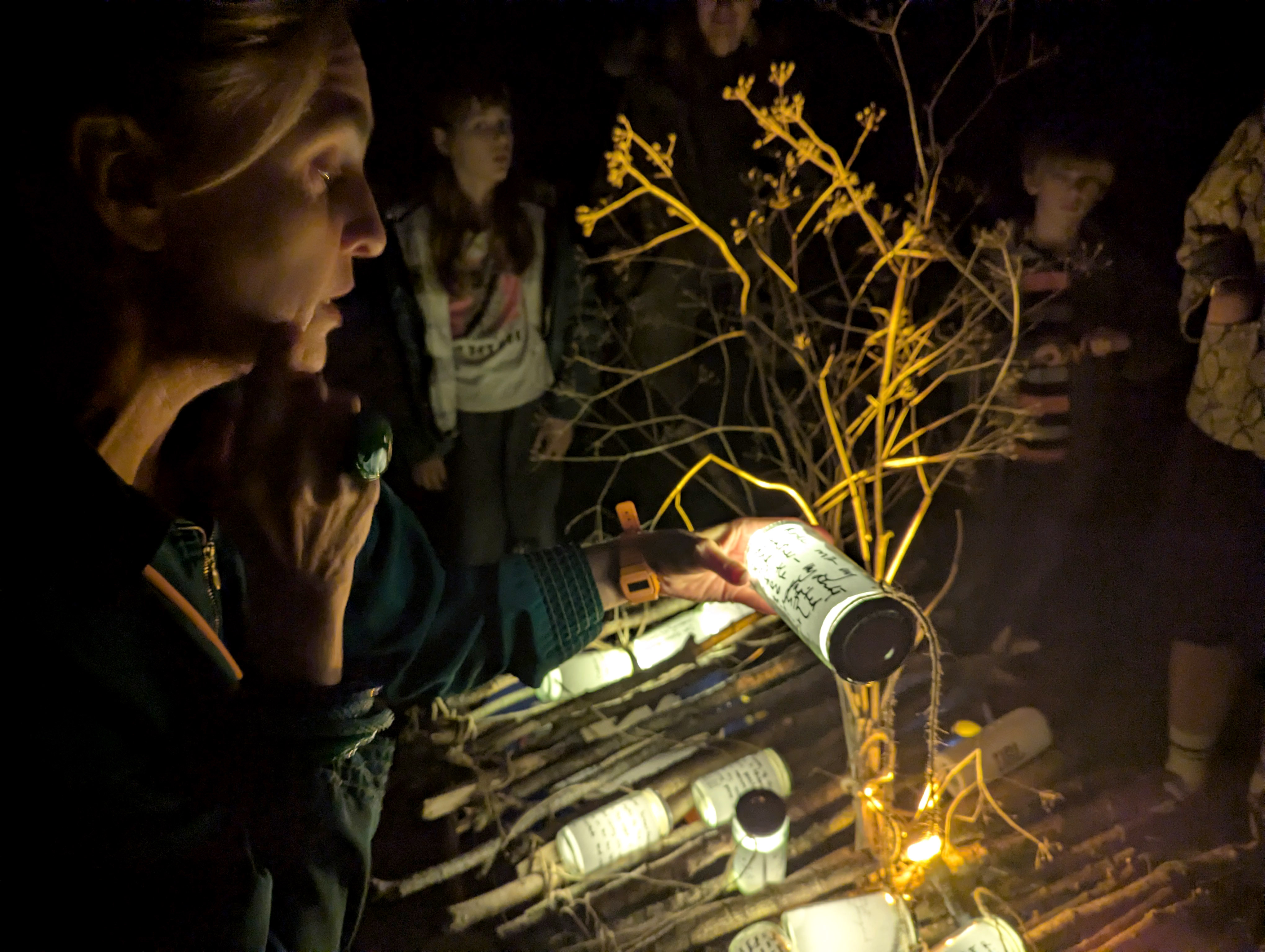 A woman holds a flashlight over a memorial display of candles, flowers and handwritten notes, with several people standing in the dark behind her.