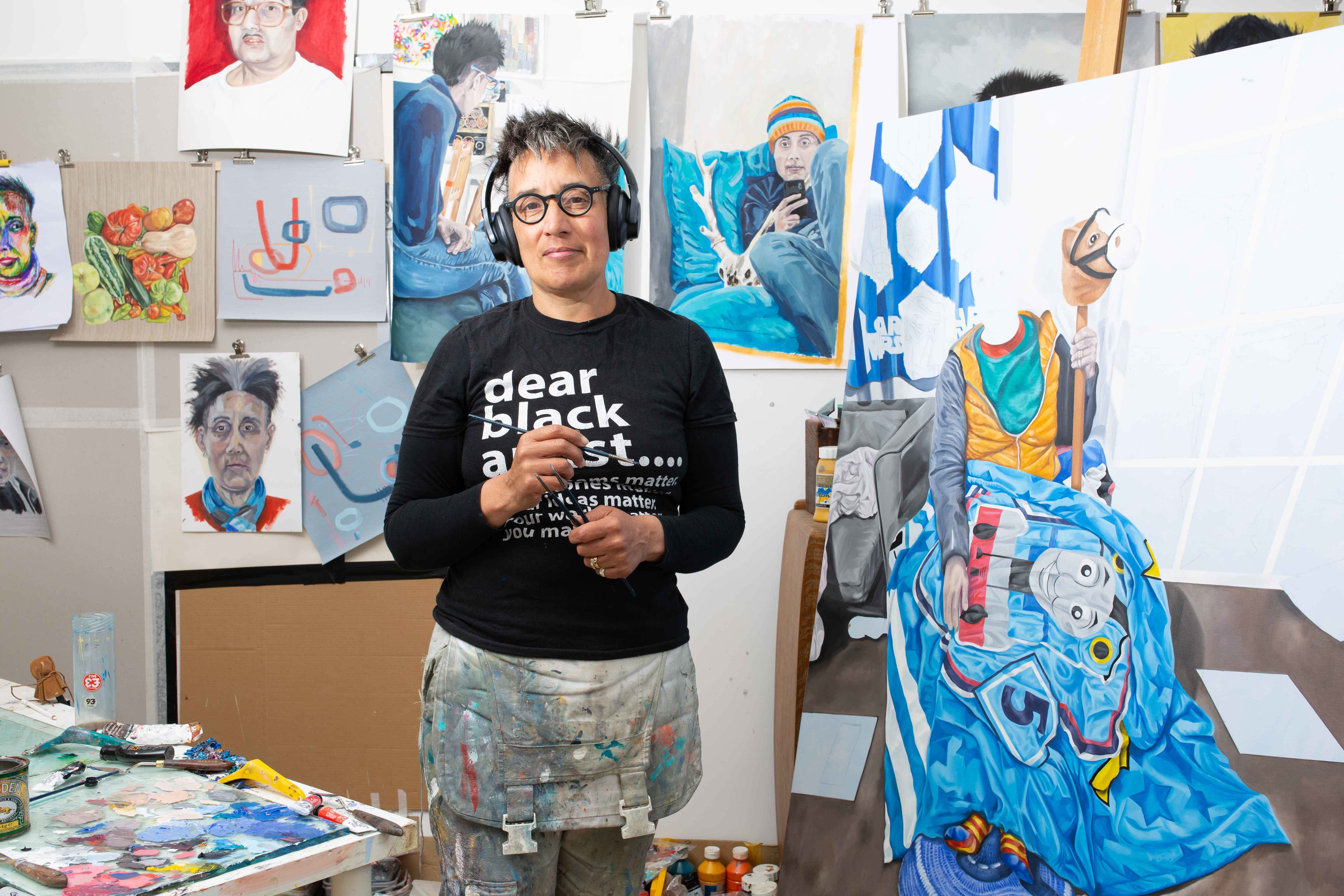 A woman poses for a portrait in an art studio, holding a paintbrush. Completed and part-completed artworks are visible to the right-hand side and in the background.
