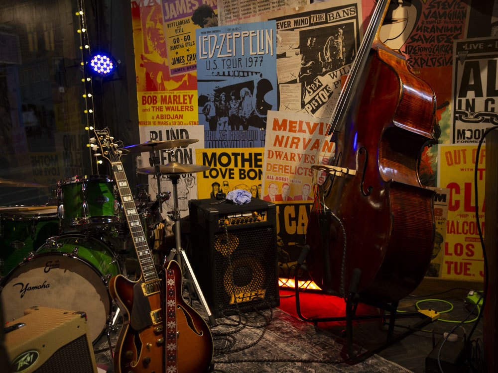 A sunburst electric guitar and upright double bass stand beside a drum kit and amplifier in a music room, with rock band posters covering the wall behind them.