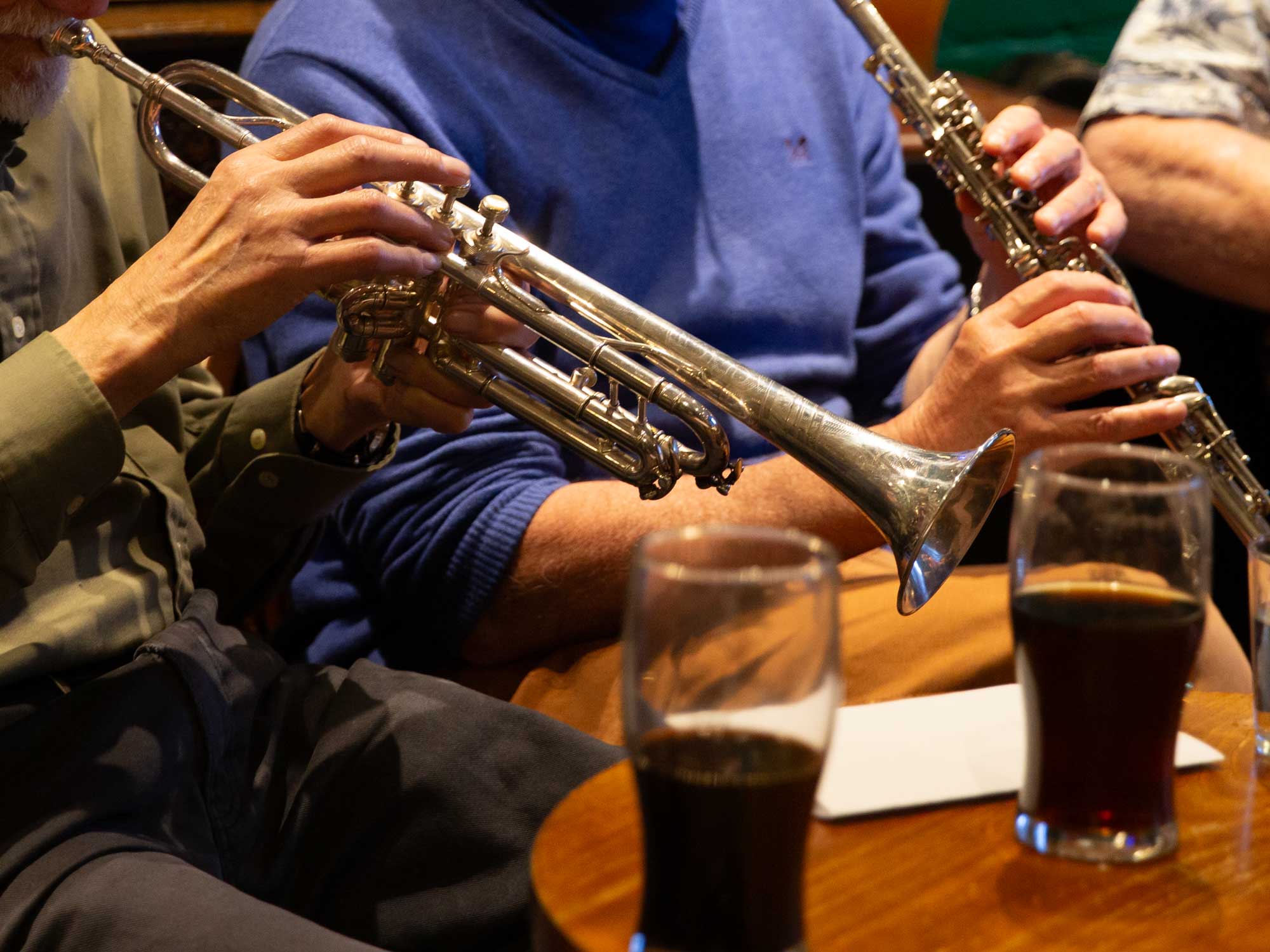 A person playing a trumpet beside another holding a clarinet at a wooden table, with two glasses of dark drink in the foreground.