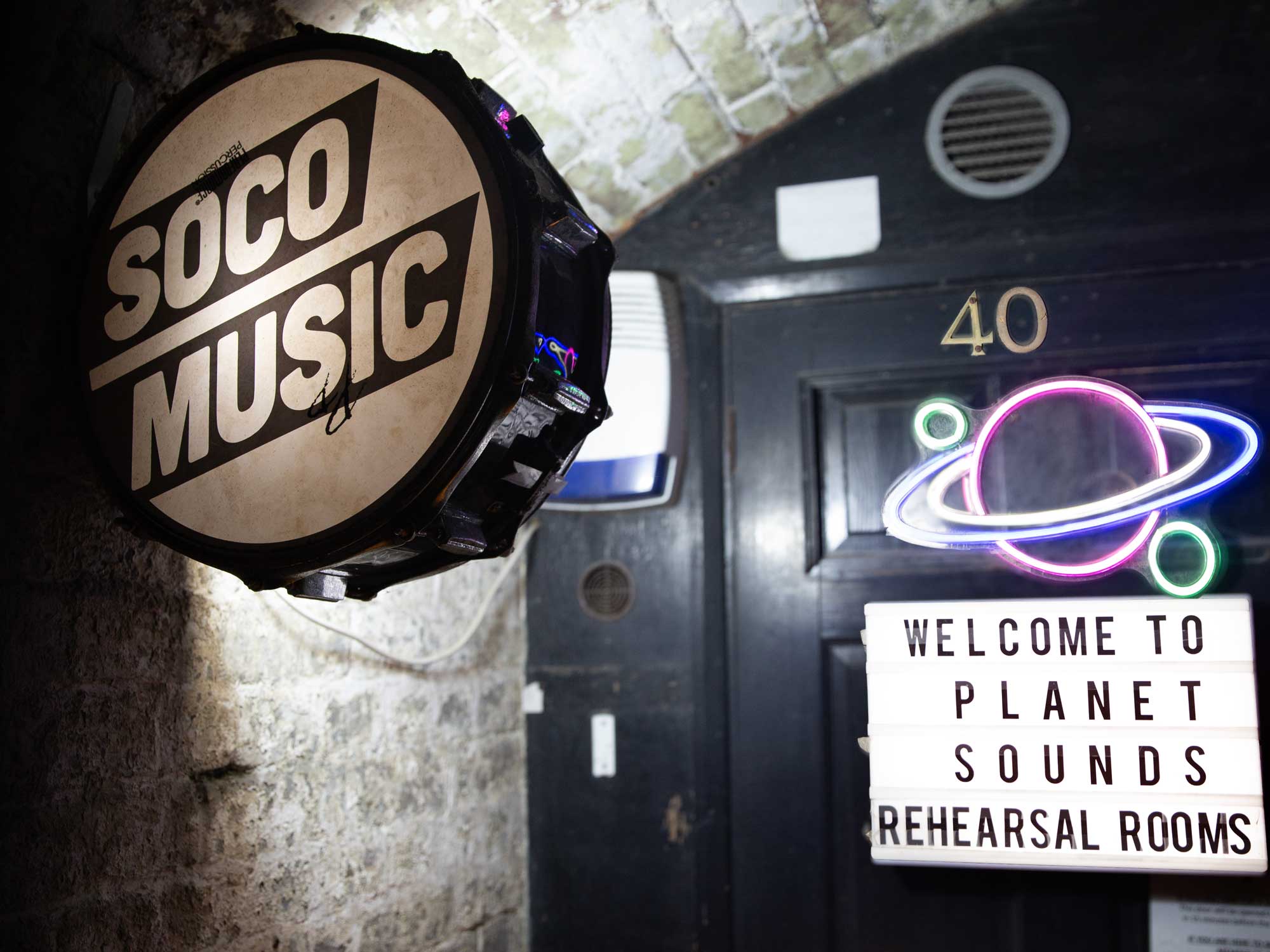 A SOCO Music drum sign hangs beside a black doorway marked 40, with a neon planet sign and a lightbox reading 'Welcome to Planet Sounds Rehearsal Rooms'.
