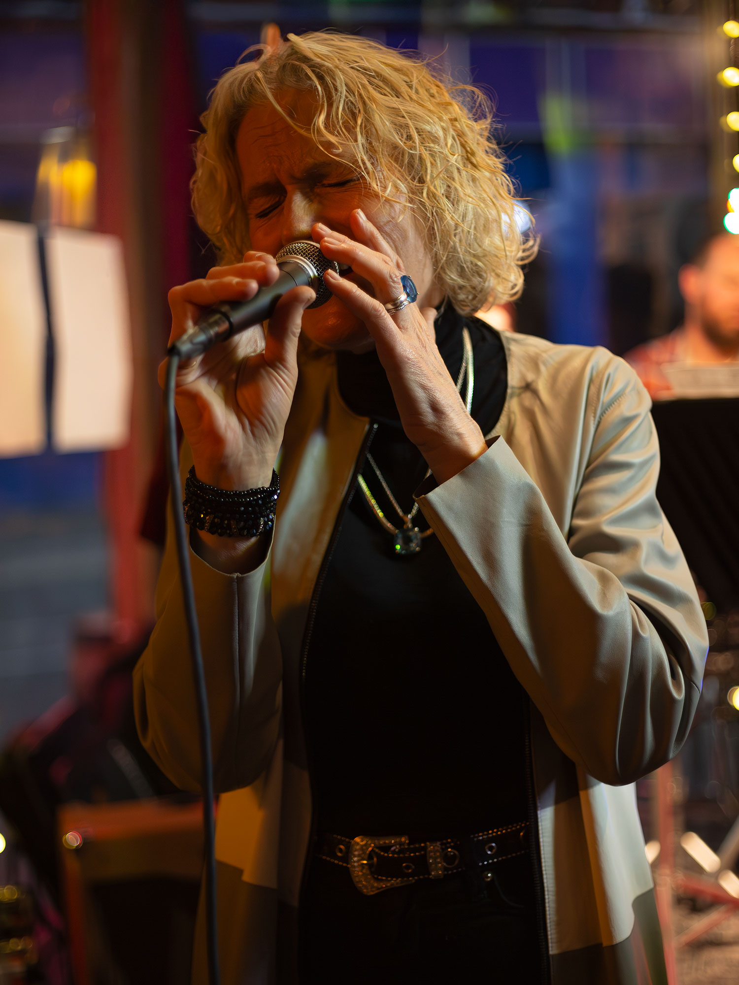 A blonde singer holds a microphone with both hands during a live performance, wearing a beige blazer over a black outfit with blurred stage lights in the background.