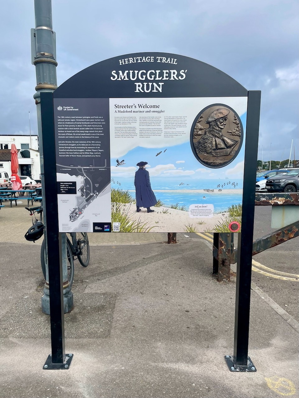 Heritage Trail information board titled "Smugglers' Run" with text, coastal illustration showing figure in blue coat, and bronze medallion.