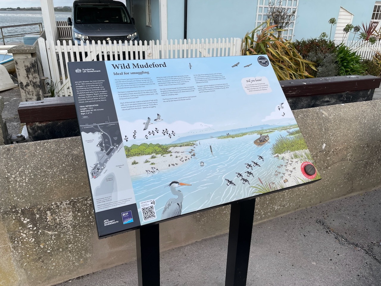 Interpretive panel titled "Wild Mudeford" showing bird illustrations and coastal map, mounted on black posts near white fence and planters.