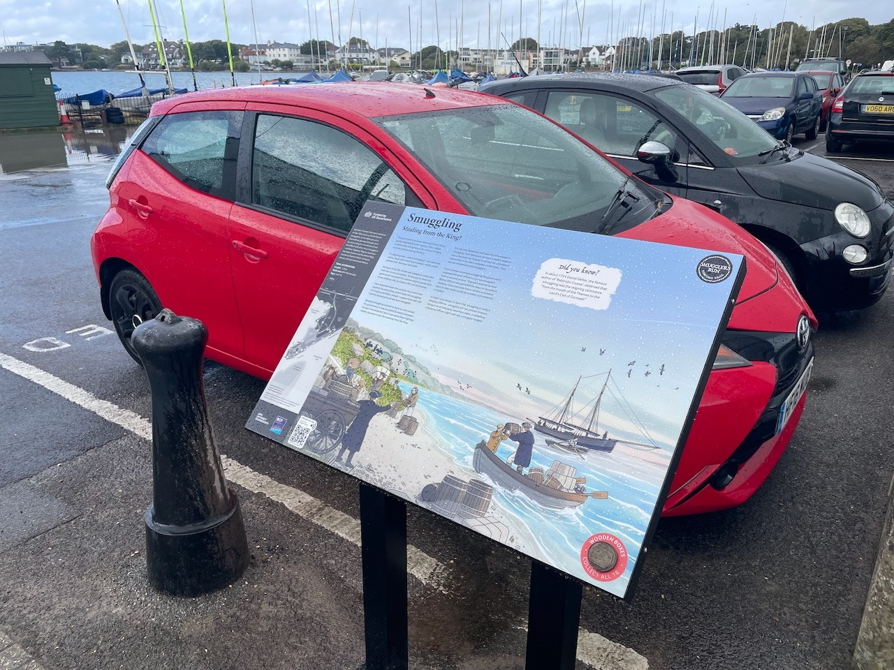 Information board in car park with red hatchback behind, marina with masts visible in background, grey cloudy sky overhead.