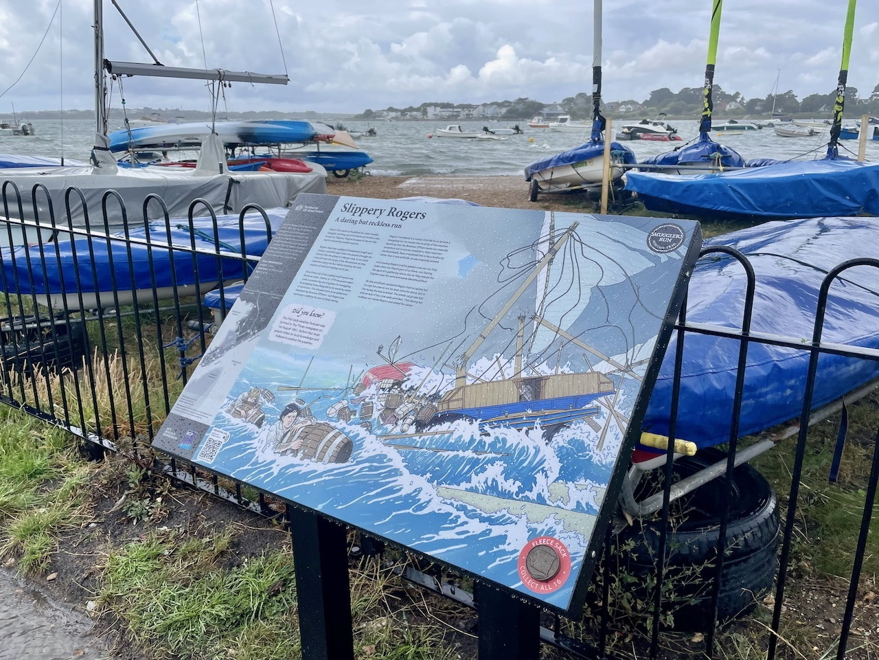 Interpretation board with map and text mounted on stand beside black railings, overlooking harbour with blue boats and cloudy sky.