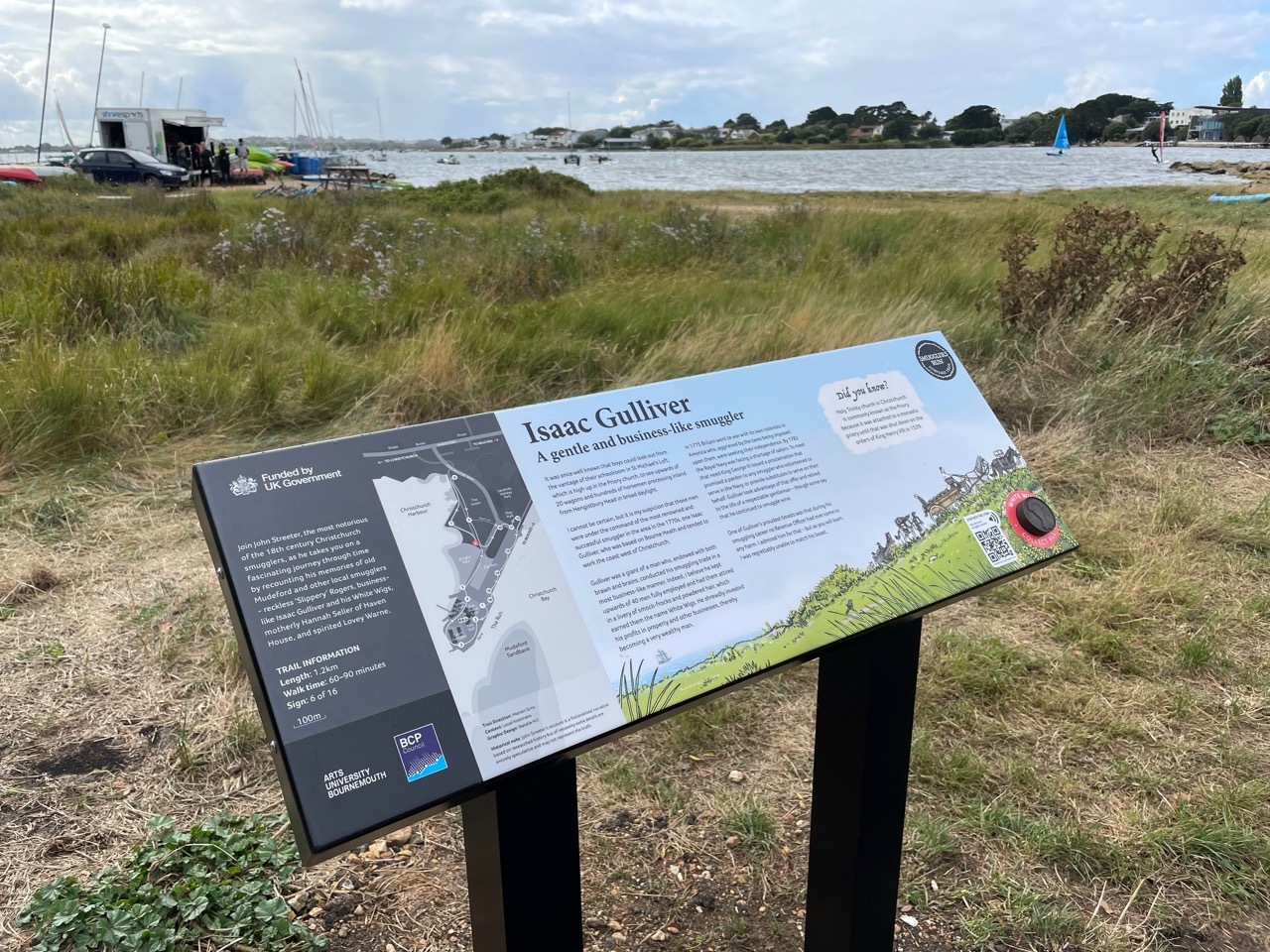 Information board titled "Isaac Gulliver" on black posts, overlooking grassy area with boats moored in harbour beyond.