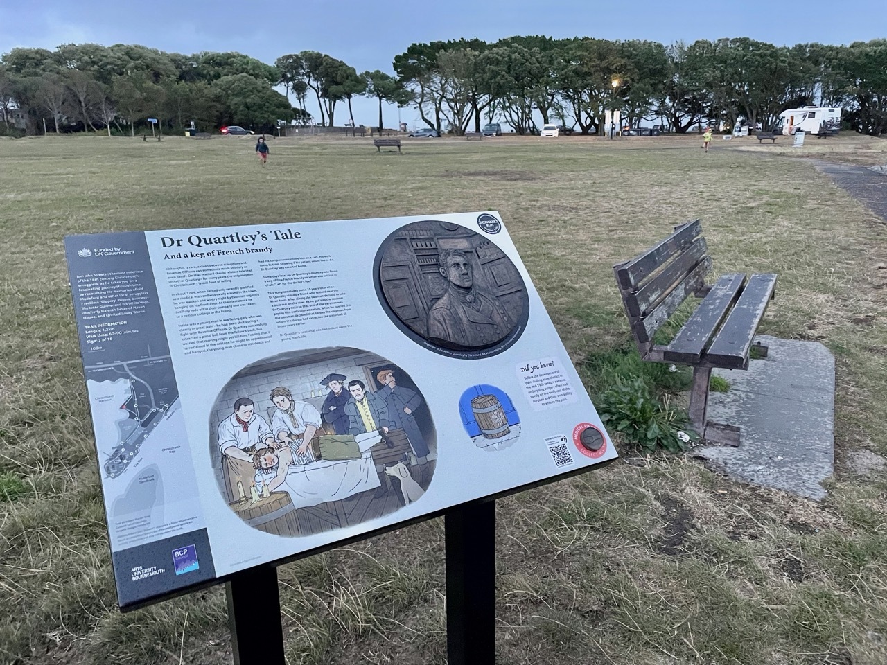 Interpretive sign about Dr Quarley's Isle with historical images and text, wooden bench behind, open grassy field with trees in background.