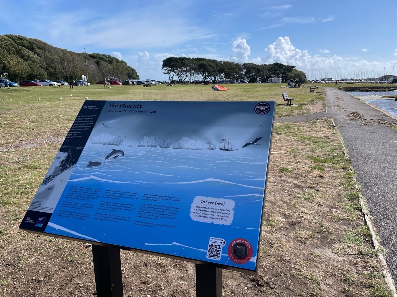 Interpretive sign with blue water scene mounted on black stand in grassy coastal area with trees and cloudy sky behind.
