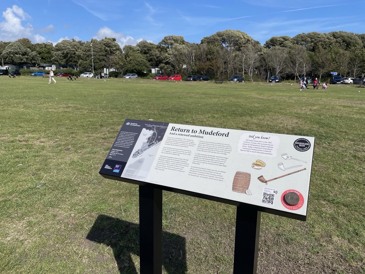Interpretive sign titled "Return to Mudford" on metal stand in large green field with trees and parked cars in background.