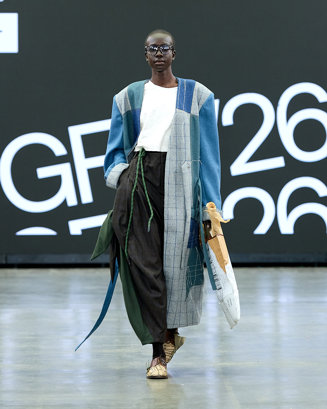 A model walks down the runway wearing a blue and grey oversized coat over a white top, paired with black trousers. The background features large digital text and a minimalist design, typical of a fashion show setting.