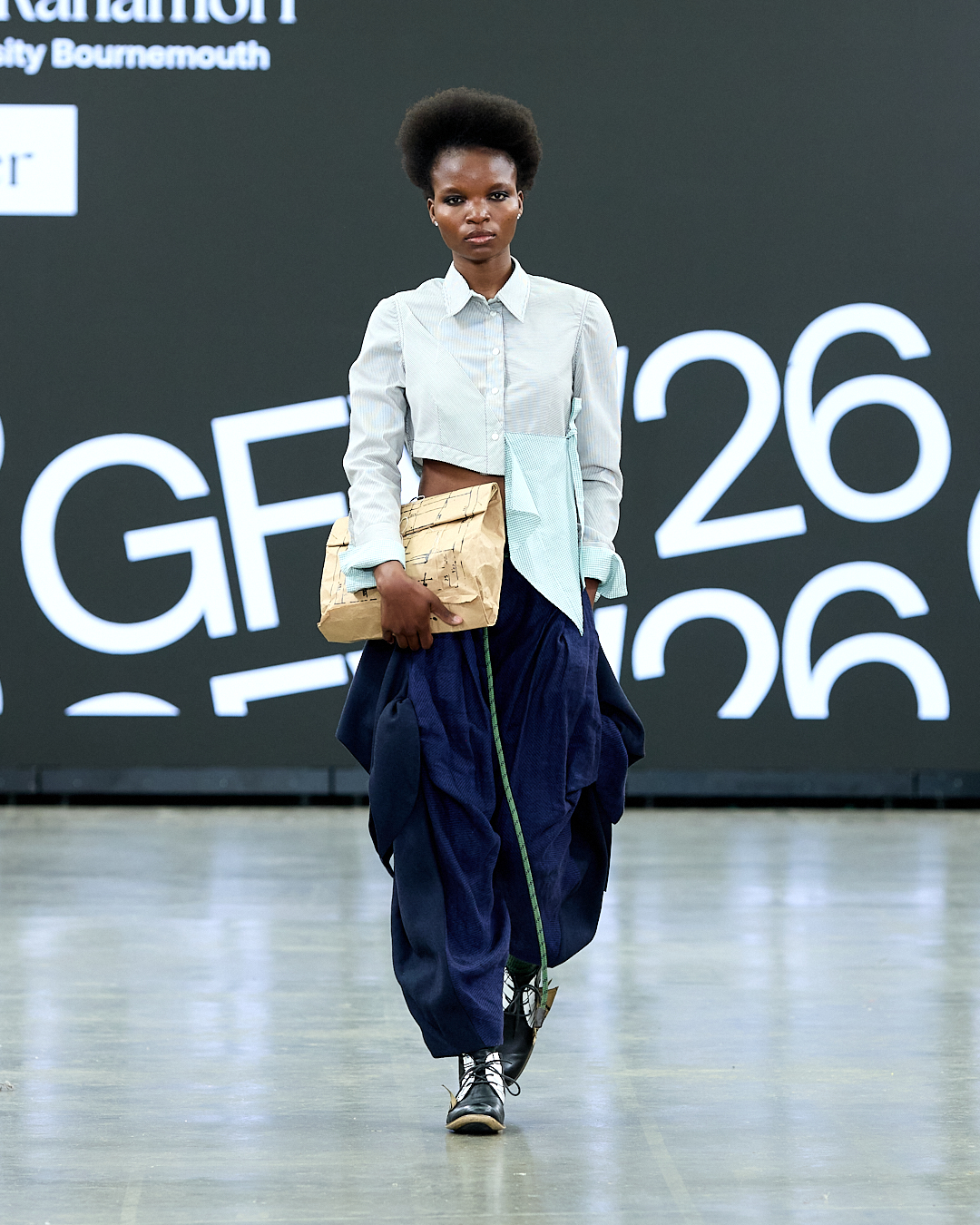 A model walks down the runway wearing a light blue and white button-up shirt paired with navy baggy trousers. She carries a crumpled gold clutch bag and wears black ankle boots. The background features large display screens.