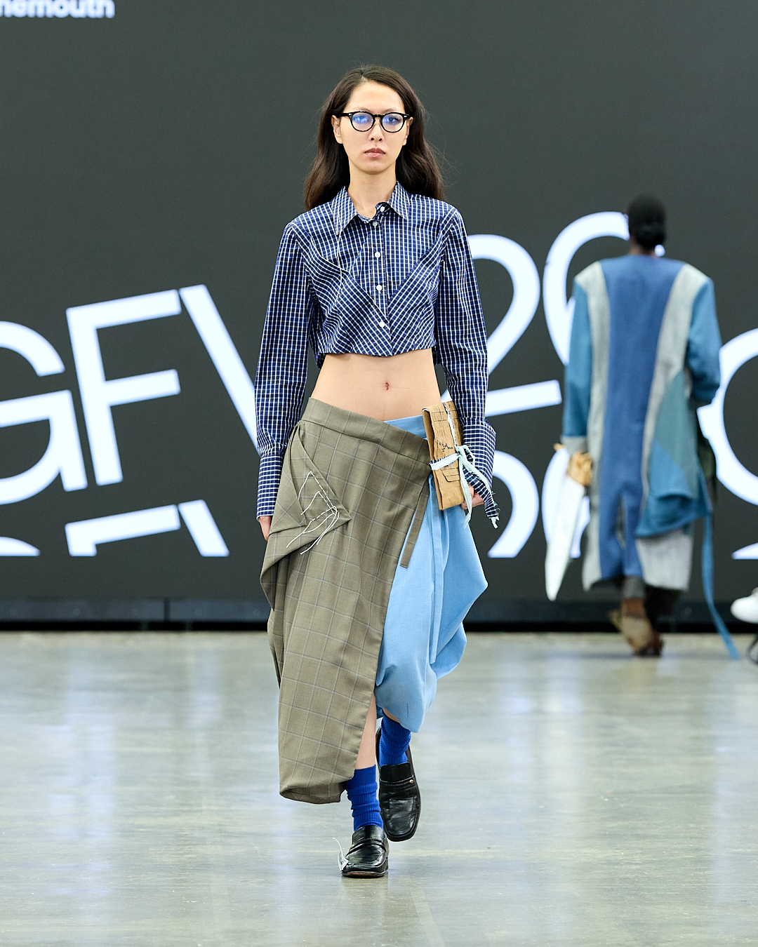 A model walks down the runway wearing a cropped blue and white checked shirt and an asymmetrical outfit made of grey and blue fabric. She accessorises with blue socks and black shoes, with a minimalist background featuring bold text.