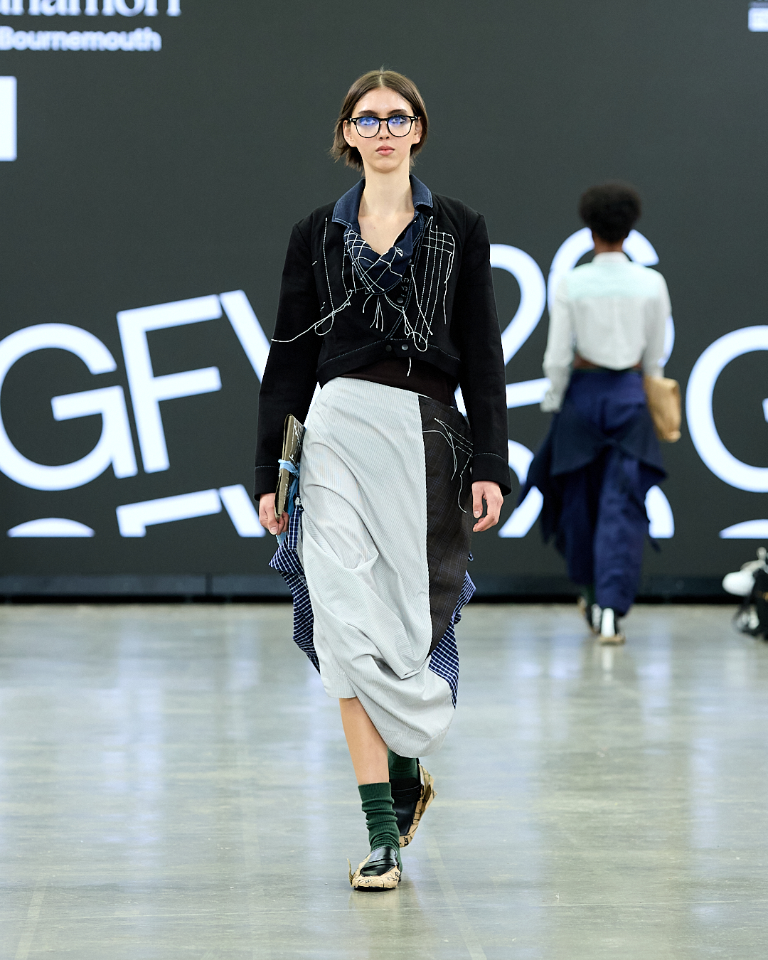 A model walks down the runway, showcasing a black blazer layered over a multi-coloured skirt featuring grey, blue, and striped patterns. The model wears glasses and green socks, with a large promotional backdrop featuring text in the background.