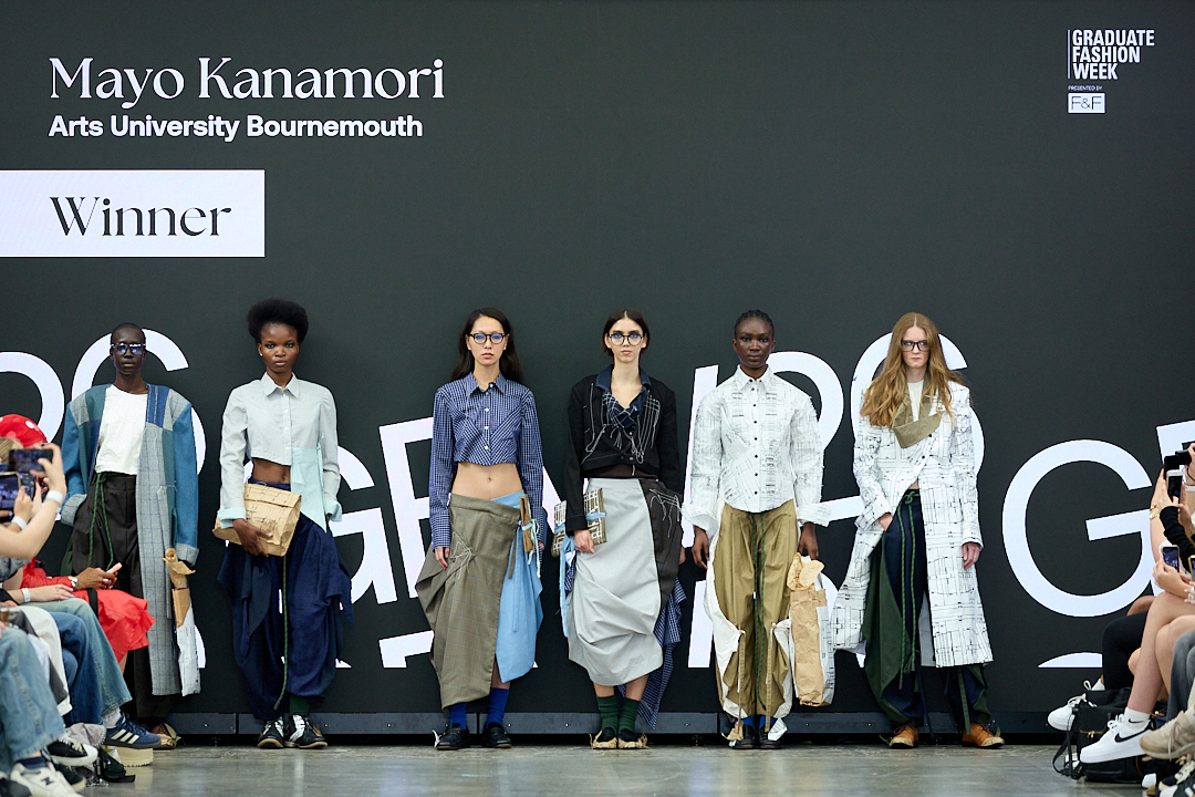 A group of six models presenting designs on a runway. They wear a mix of elevated fashion pieces, with varying styles and fabrics. The backdrop features text indicating "Mayo Kanamori" and "Winner" from the Graduate Fashion Week.