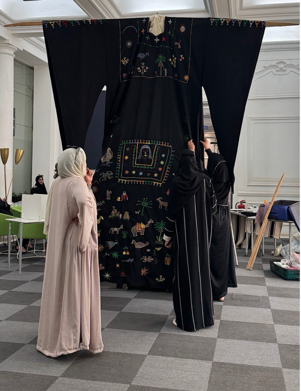 Two people in long robes examining large black garment with colourful embroidered patterns displayed in white gallery space.