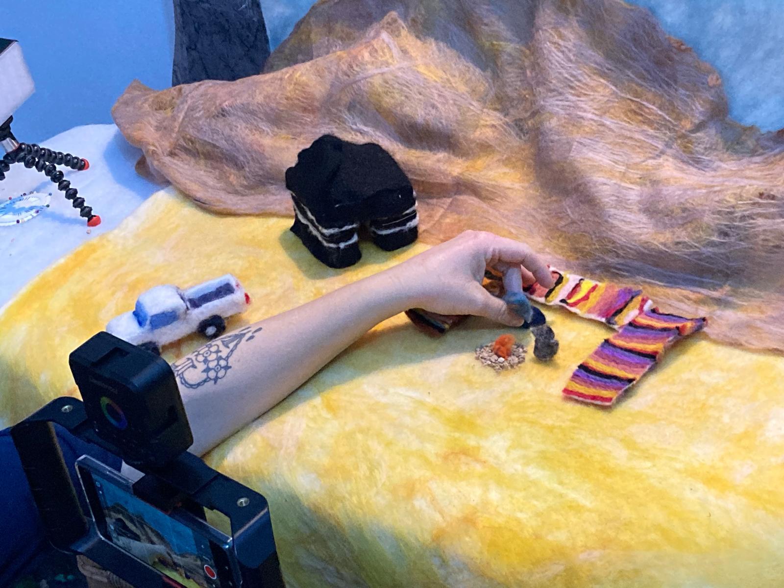 Person's hand reaching across yellow surface towards striped object, with black equipment and beige fabric in background.