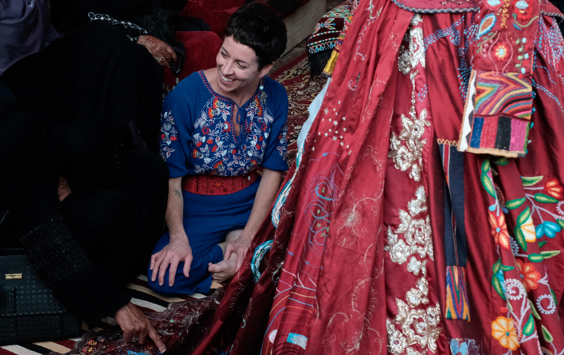 Person in blue embroidered shirt with red sash sits amongst colourful traditional garments including red floral dress and patterned fabrics.