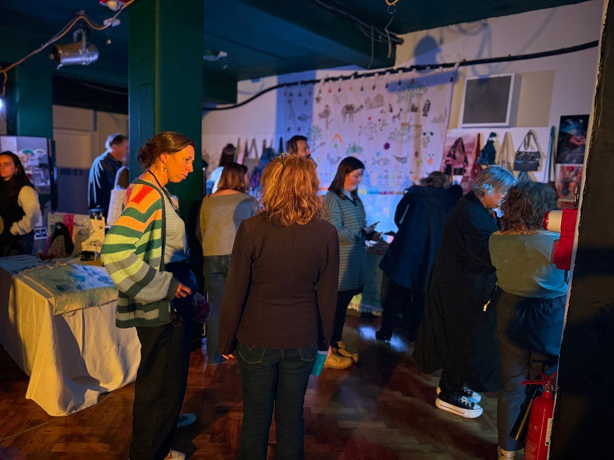 Group of people gathered around white display tables in dimly lit indoor space with wall exhibits and coloured lighting.