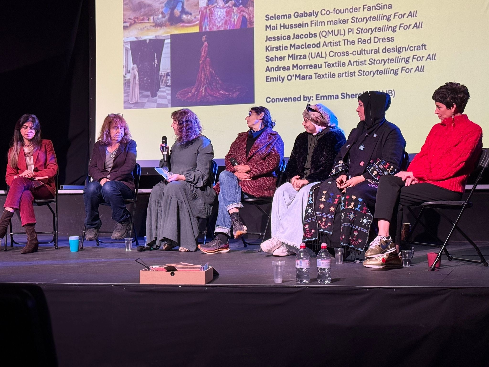 Seven people seated on chairs on a stage with projection screen showing text and images behind them. Panel discussion setup.