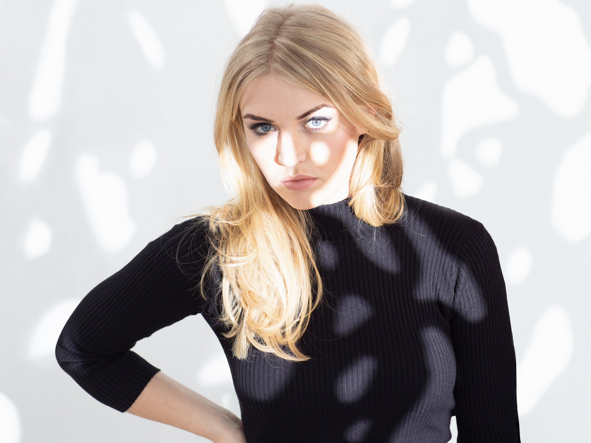 Blonde woman in black ribbed top with hand on hip against white geometric patterned background.