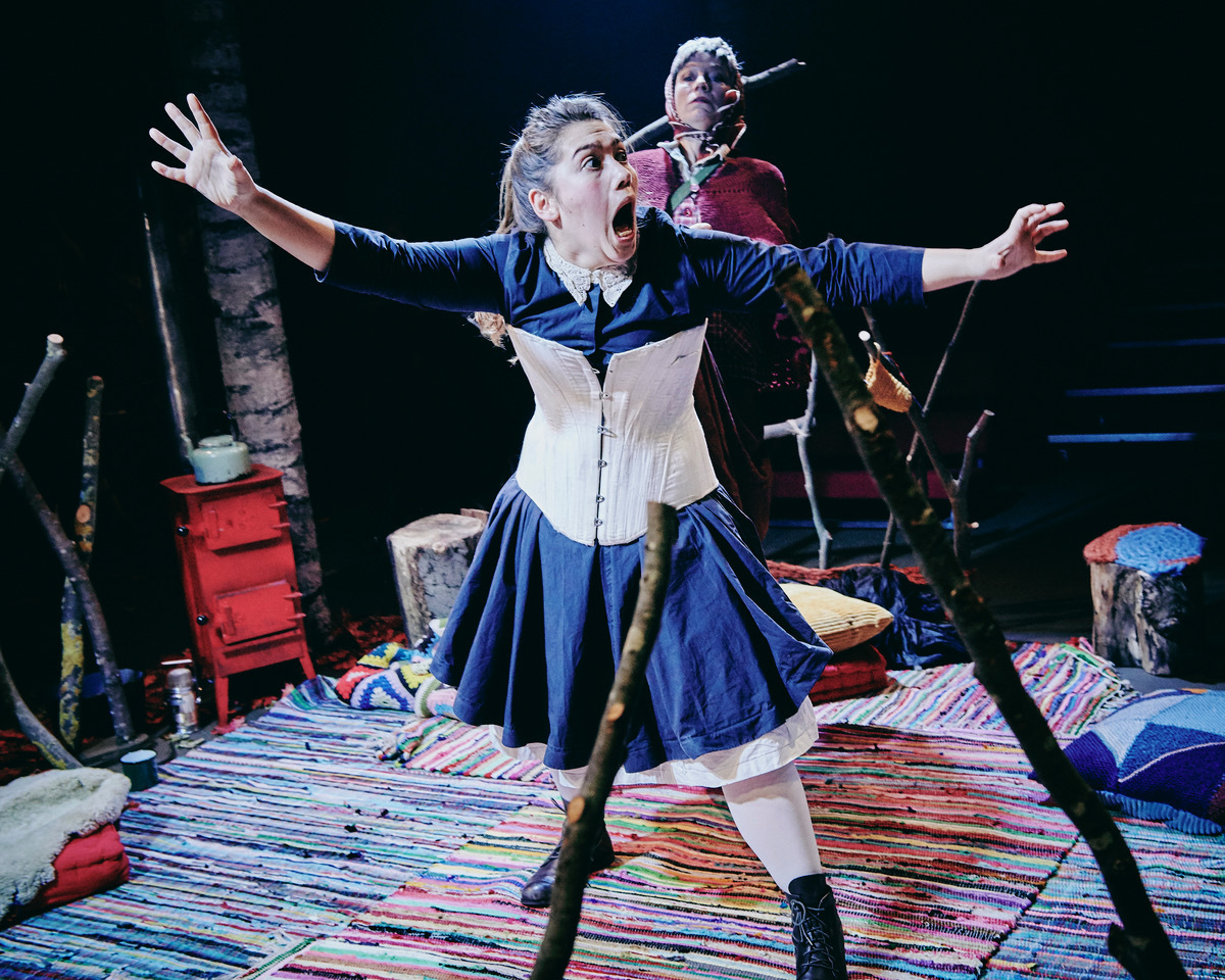 Woman in blue dress and white corset kneels on striped rug with arms outstretched, mouth open. Man with microphone stands behind her.