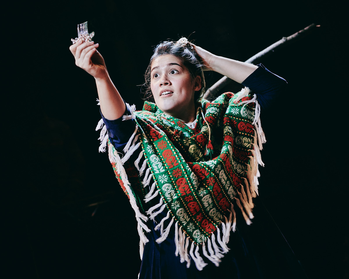 Woman on stage holding microphone and crystal trophy, wearing green patterned poncho with white fringe against black background.
